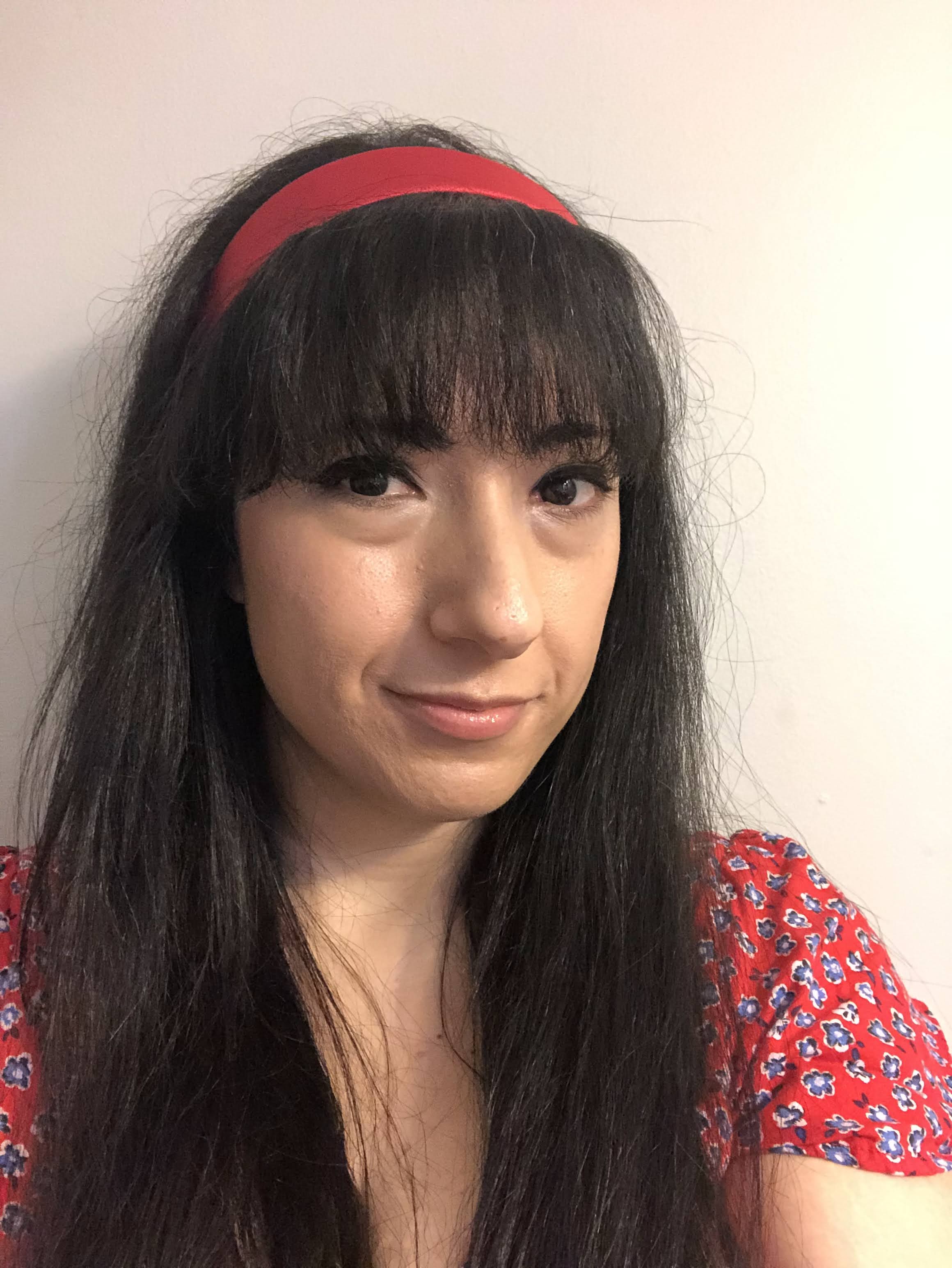 A close-up portrait of an Arab Woman with long black hair and bangs, wearing a red headband and a red floral top, smiling softly against a neutral wall.